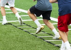 An agility ladder is on a green turf field with three young boys in cleats running through each leg.