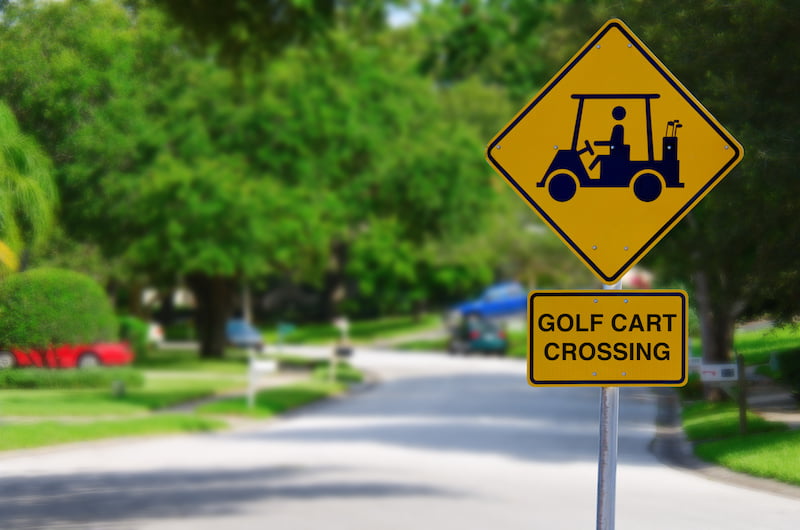 golf cart crossing sign on a residential street intersection with blurred lush green trees in the background.