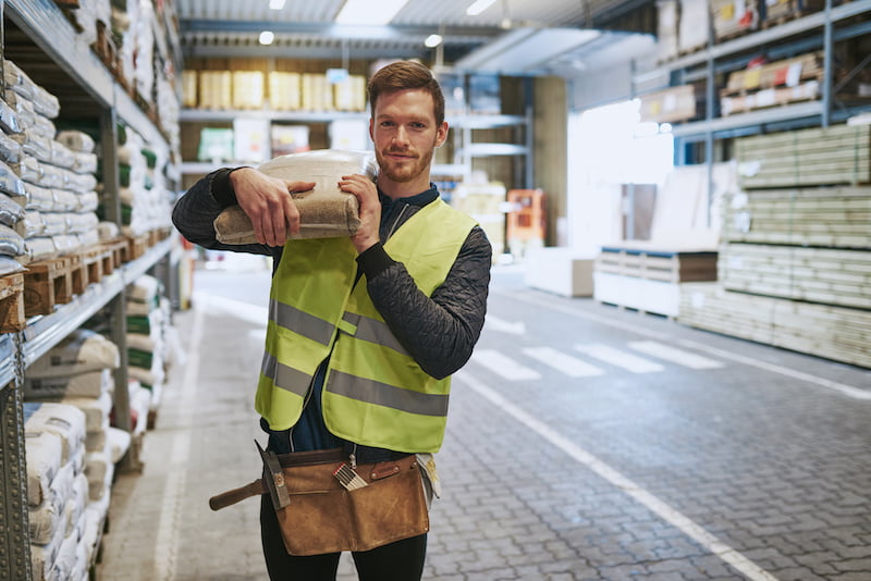 young handyman selecting a pocket of product in a warehouse standing with the bag over his shoulder smiling at the camera