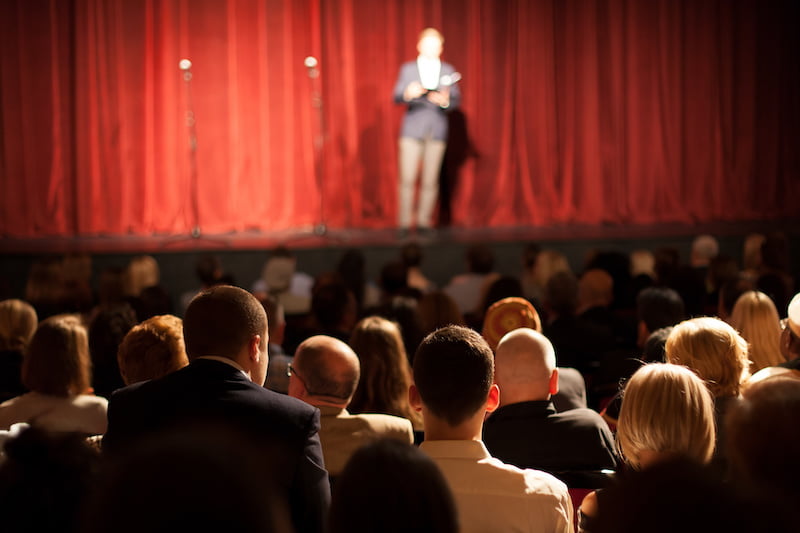 comedian on stage in front of red curtain, large audience.