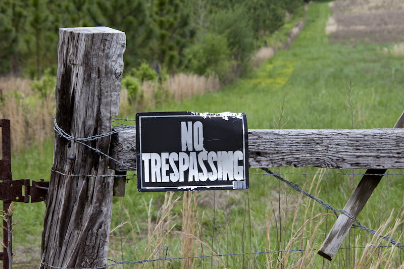 warning sign for no trespassing on an old, weathered fence post with barbed wire at the entrance of a green meadow.