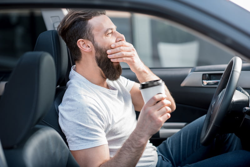 tired man yawning on the front seat of the car holding coffee to go.