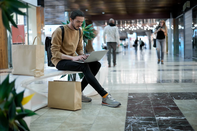 man sitting on bench, using computer after shopping with friends.