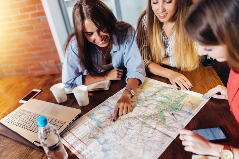 smiling female friends sitting at desk planning their vacation looking for destinations on map.