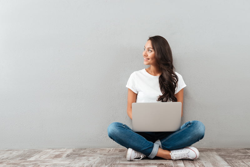 attractive asian woman holding laptop computer while sitting on the floor with legs crossed and looking away at copy space isolated over gray background