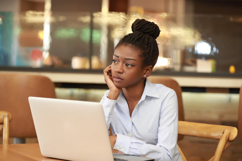young beautiful african girl sitting at table with laptop leaning on hand and thinking.