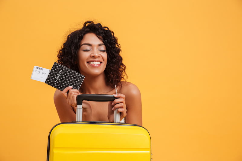 portrait of a smiling young african woman dressed in summer clothes holding passport with flying tickets while standing with a suitcase isolated over yellow background
