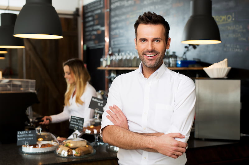 happy small business owner standing at front of bar with employee in background preparing coffee.