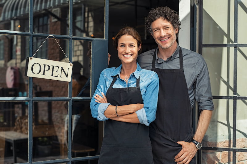 two cheerful small business owners smiling and looking at camera while standing at entrance door. happy mature man and mid woman at entrance of newly opened restaurant with open sign board. smiling couple welcoming customers to small business shop.