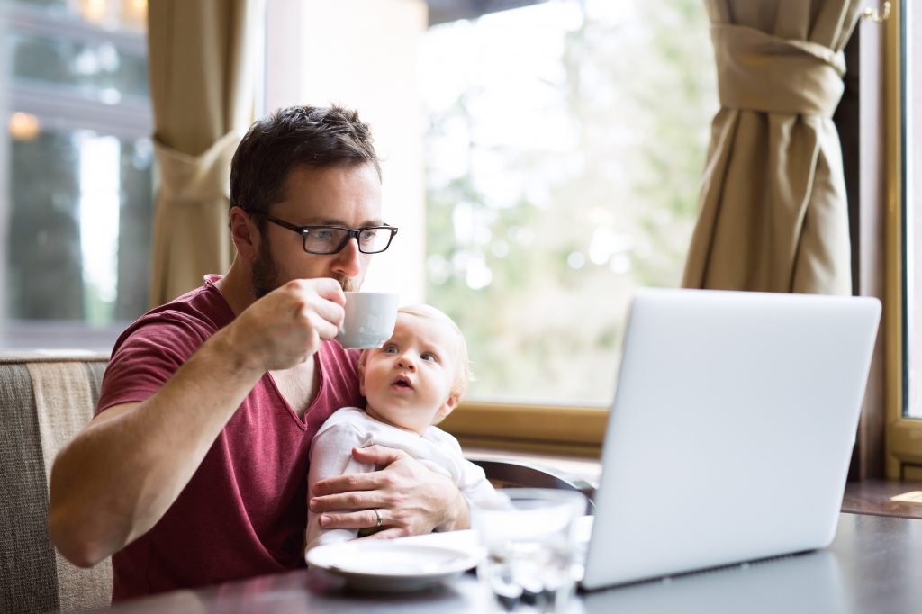young handsome man with notebook in cafe sitting at the table drinking coffee, holding his son in his lap