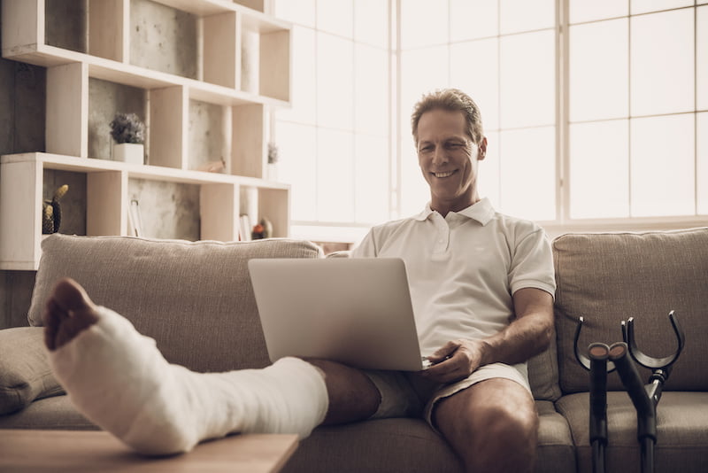 man with fractured leg sit on sofa and use laptop.