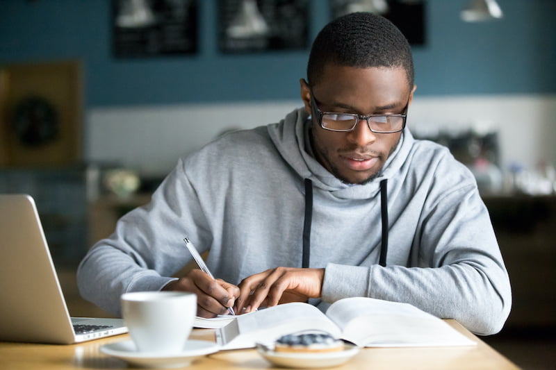 focused millennial student in glasses making notes writing down information from book